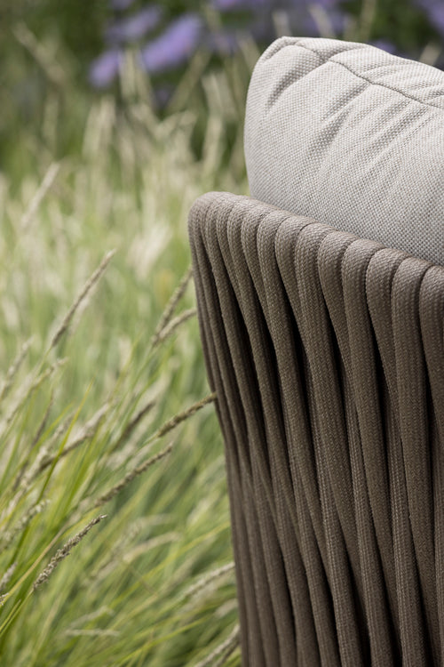The image features the back of a stylish outdoor chair with a ribbed design, showcasing a soft, beige cushion resting on top. Surrounding the chair are tall, wispy grasses, creating a serene garden atmosphere.