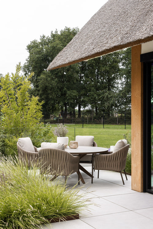 A casual outdoor dining area features a round low dining table surrounded by four stylish chairs with toffee-colored cushions. The setting is complemented by lush green landscaping and a thatched roof structure, creating a serene ambiance.