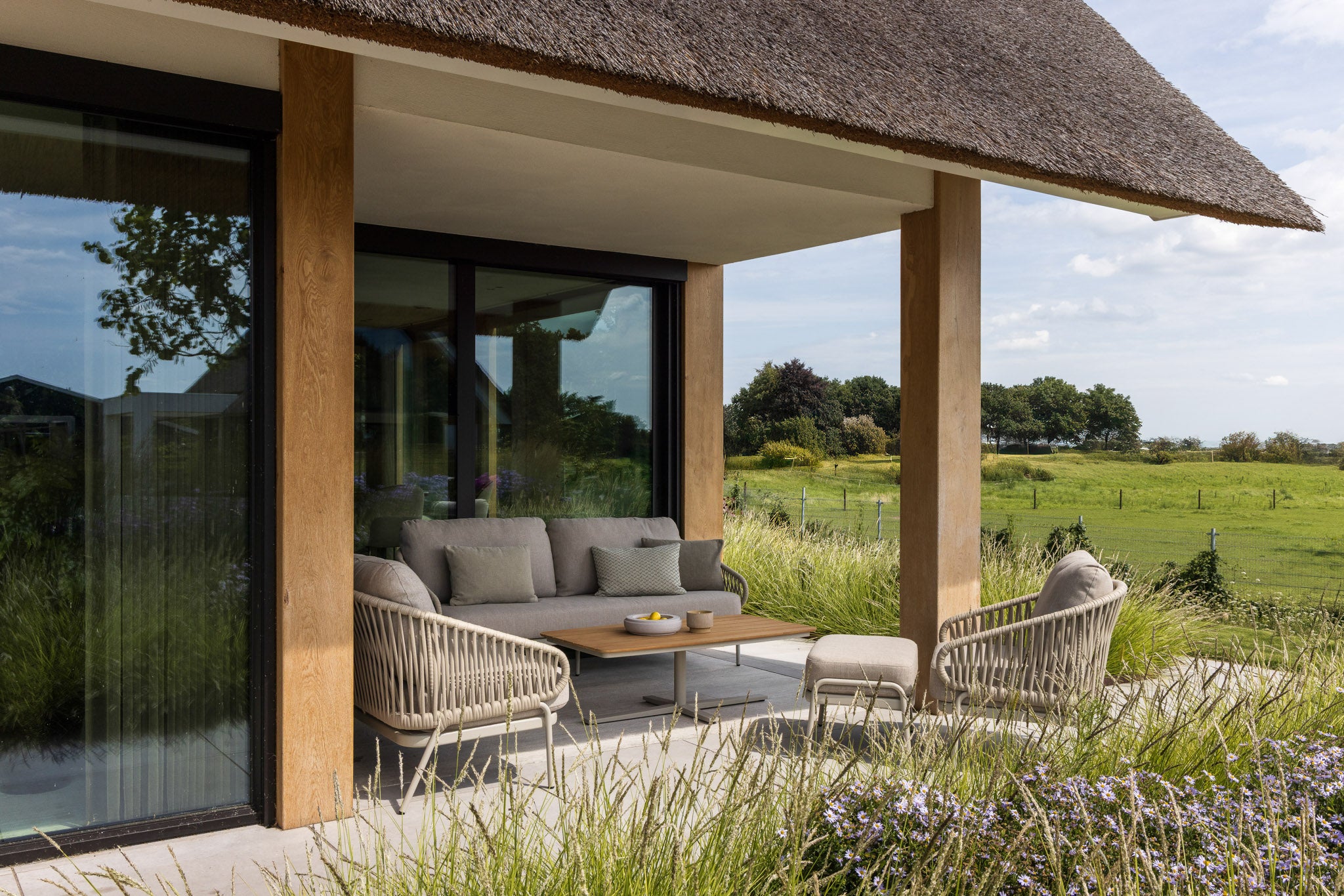 Modern outdoor seating area with a thatched roof and glass walls, overlooking a green landscape.