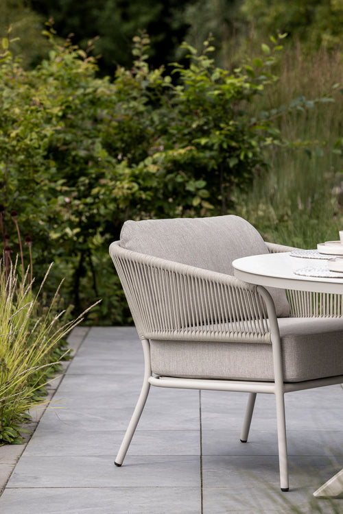 A close-up view of a light beige outdoor dining chair with a woven backrest, positioned beside a round dining table on a stone patio. Lush greenery and plants frame the scene, creating a tranquil outdoor dining atmosphere.
