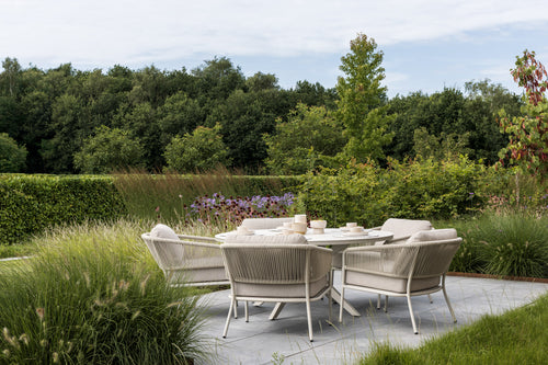 A stylish outdoor dining set featuring a round table surrounded by five beige cushioned chairs, set against a lush green backdrop of trees and flowering plants. The serene atmosphere is enhanced by soft grass and colorful foliage.