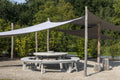 Outdoor setting with a large shade sail over a round table and chairs, surrounded by greenery.