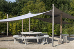 Outdoor setting with a large shade sail over a round table and chairs, surrounded by greenery.
