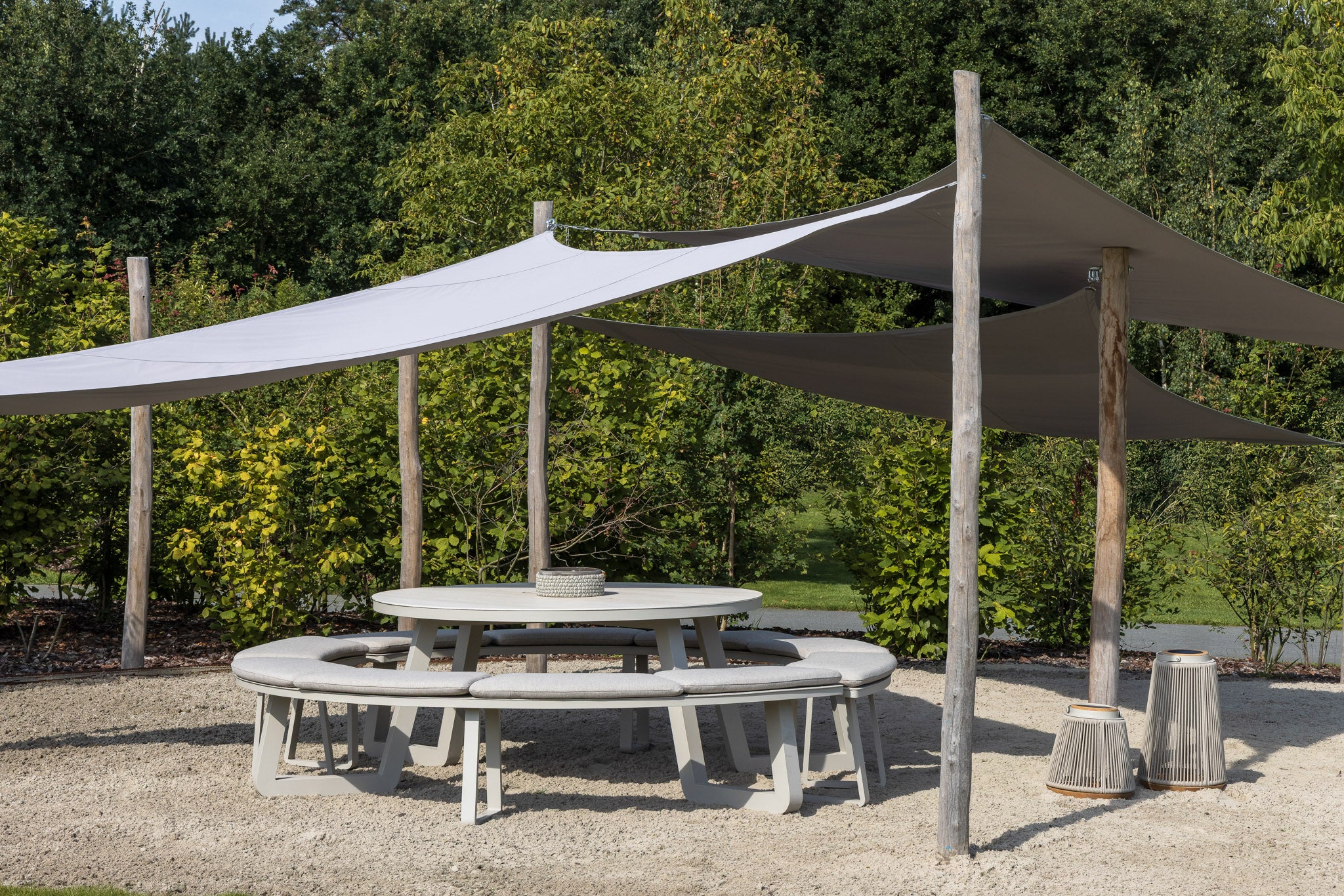 Outdoor setting with a large shade sail over a round table and chairs, surrounded by greenery.