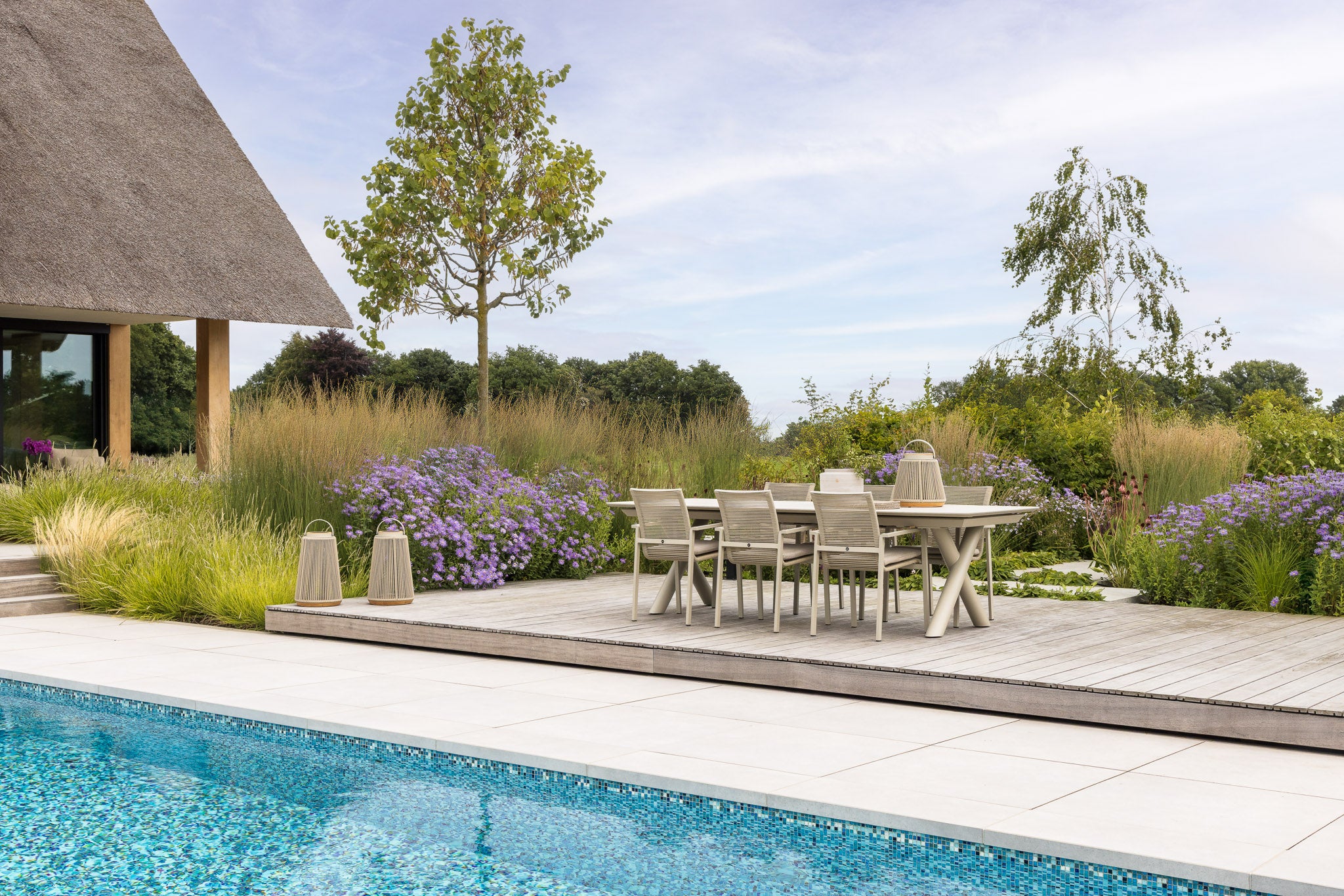 Outdoor dining area with table and chairs near a pool, surrounded by greenery and flowers.