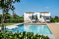 Modern outdoor poolside scene with a rectangular swimming pool, white lounge chairs, two beige umbrellas, a white wall structure, green landscaping, and a clear blue sky.