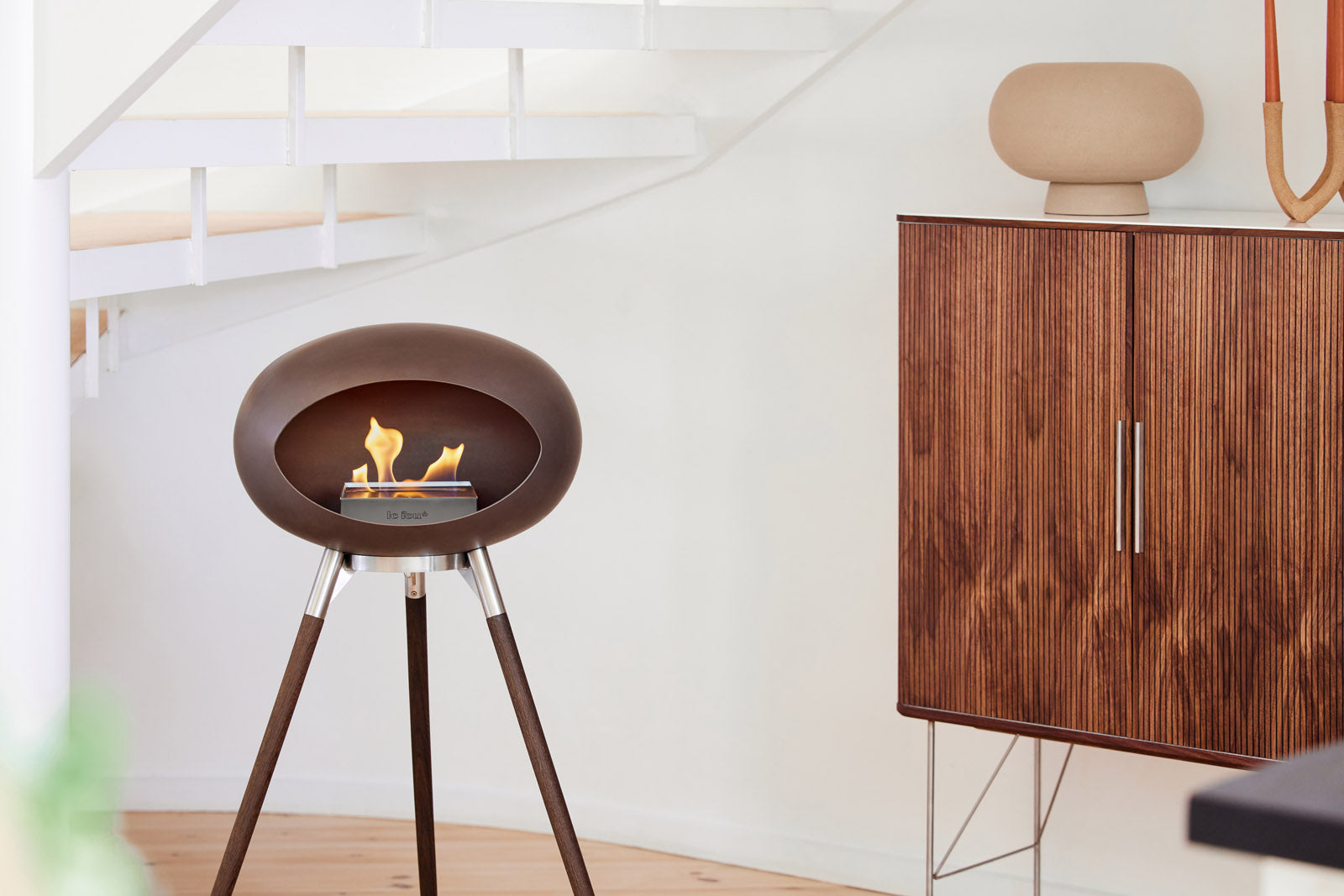 A modern oval freestanding fireplace with a dark brown finish and tripod legs, shown lit with a small flame in a minimalist interior setting next to a wooden cabinet.