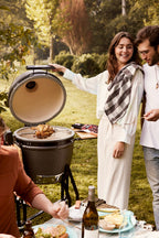 People enjoying a barbecue outdoors with a woman opening a grill and a man holding a bottle.