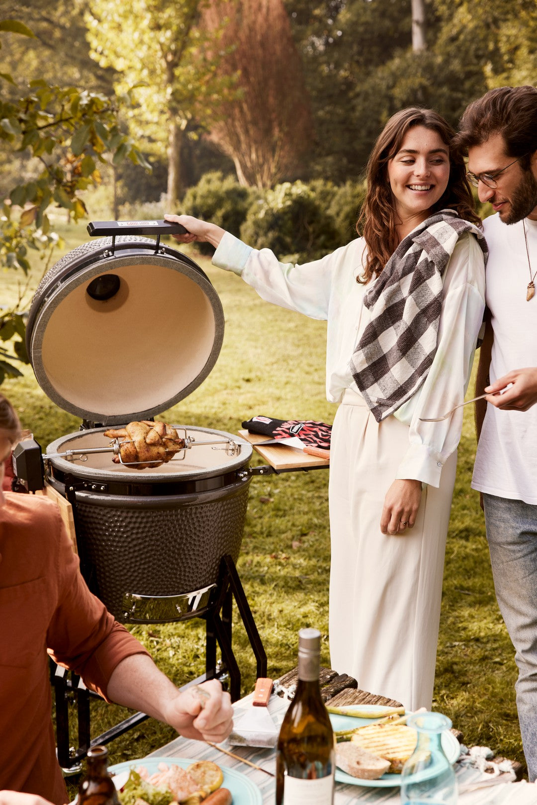 People enjoying a barbecue outdoors with a woman opening a grill and a man holding a bottle.