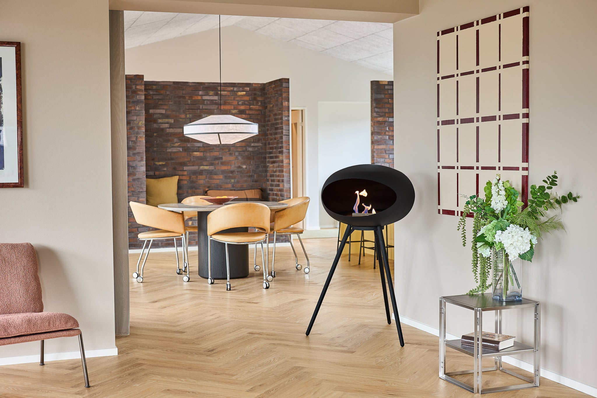 A modern black oval freestanding fireplace or fire pit with a small visible flame, placed in a styled interior room with wood flooring, dining furniture, and wall decor.