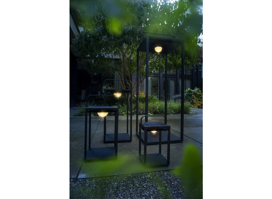 Three illuminated outdoor lanterns of different heights displayed in an evening garden setting with greenery and a building in the background
